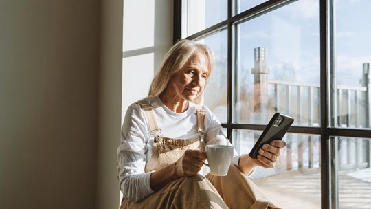 Older woman sitting next to a window checking her smartphone and drinking coffee