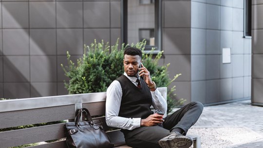 Young businessman sitting on a bench outside, talking on a phone