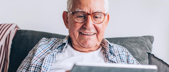 A close up of an old man in his 70s sitting on the couch at home, searching the Internet on his digital tablet, enjoying