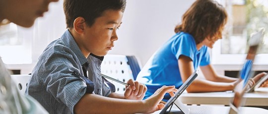 Focused Asian school boy using digital tablet at class in classroom. Attentive junior school student learning online virtual education digital program app tech during stem computer science lesson.