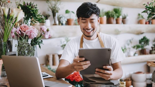 Asian male florist, owner of small business flower shop, using digital tablet while working on laptop against flowers and plants