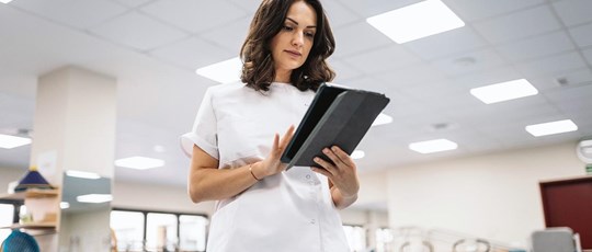 Low angle of brunette in medical uniform browsing data on digital tablet while working in spacious physiotherapy room of modern clinic