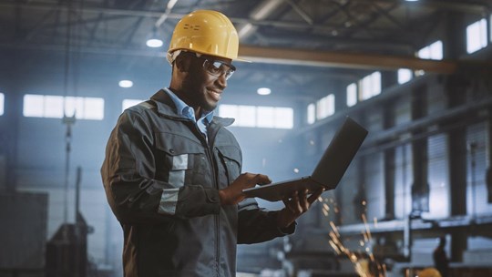Professional Heavy Industry Engineer Worker Wearing Safety Uniform and Hard Hat Uses Laptop Computer. Smiling African American Industrial Specialist Standing in a Metal Construction Manufacture.