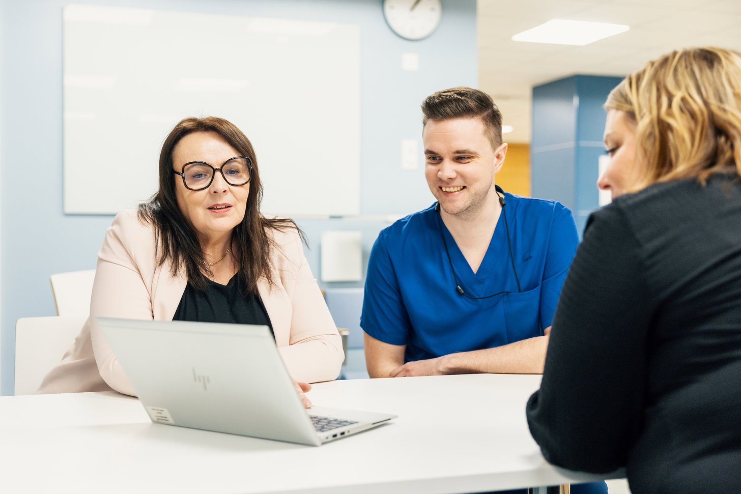 Three cheerful professionals sitting at a table looking at a laptop. From left: Marjo Patana (light pink blazer), Sami Penttil&auml; (blue nurse shirt), and Tytti Valli (black jacket). In the background: light blue walls, a hallway, and a wall clock.